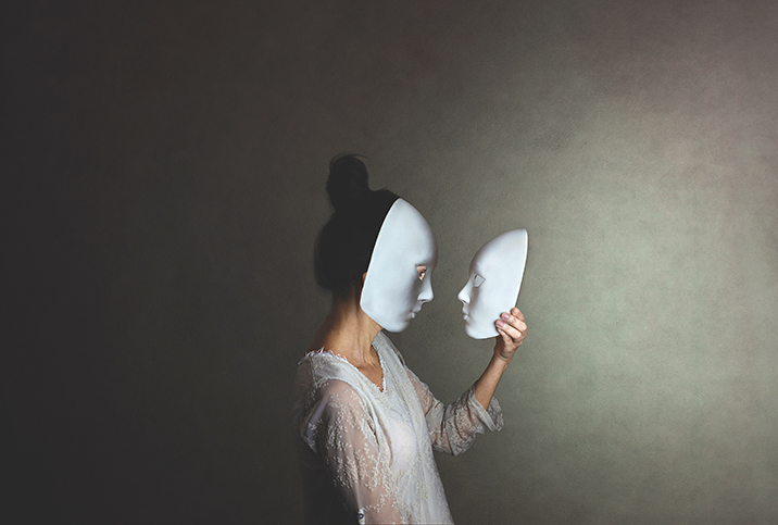 A woman in a white mask holds another mask in her hand and looks at it face to face.