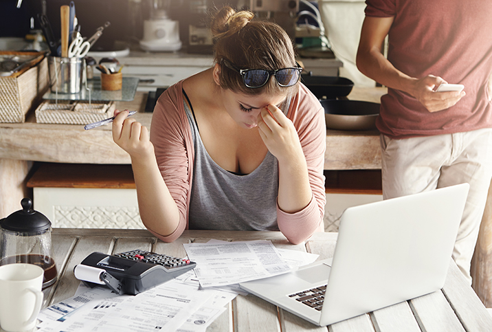 A woman holding a pen in one hand tries to concentrate and rests her forehead in her other hand, a laptop, calculator, and paper on the table in front of her. 