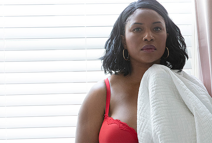 A woman in a red bra holds a white blanket and stands in front of a window with drawn blinds.