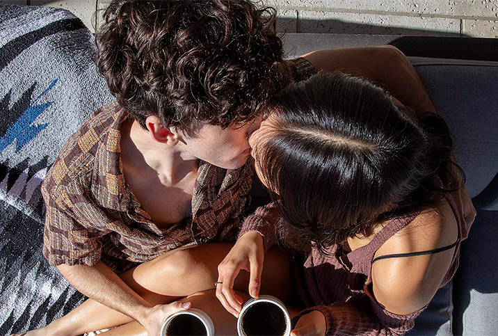 A man and and woman kiss while sitting down with cups of coffee in their hands.