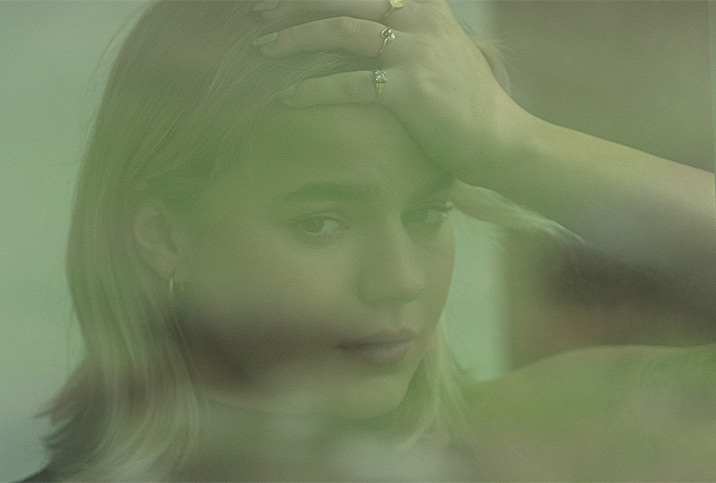 A woman looks out a green-tinted window and presses her hand to her forehead.