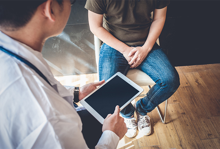 A man sits while a doctor stands in front of him holding a tablet.