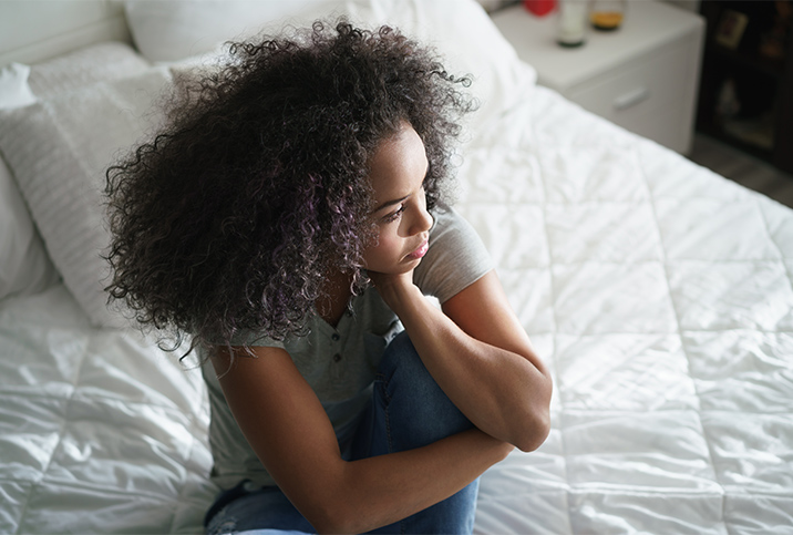 A woman sits on her bed with her leg curled up, holding her head with her hand.