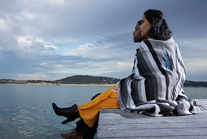 A married couple sits on the edge of a wooden dock looking out over a lake with a blanket around them.