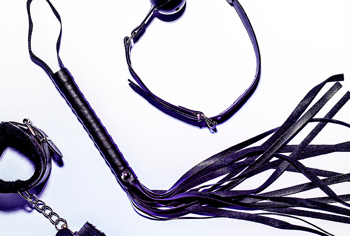 A leather whip lays between a pair of bondage cuffs and a ball gag.