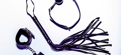 A leather whip lays between a pair of bondage cuffs and a ball gag.