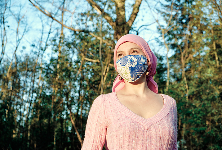 A woman in a cloth mask and head wrap stands in the sun with trees behind her.