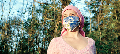 A woman in a cloth mask and head wrap stands in the sun with trees behind her.