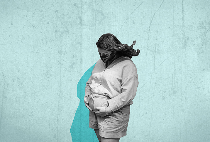 A pregnant person wears a medical mask and holds their pregnant belly with a blue background behind them.