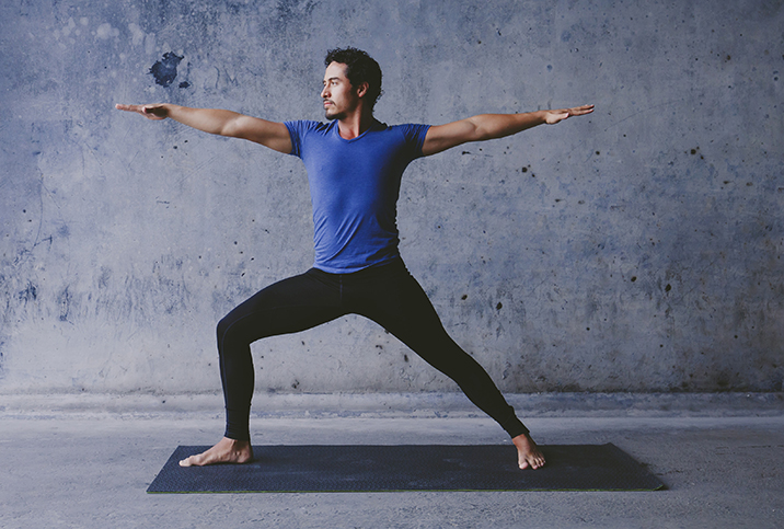 A man in black pants and a blue shirt does the Warrior II yoga pose on a black mat.