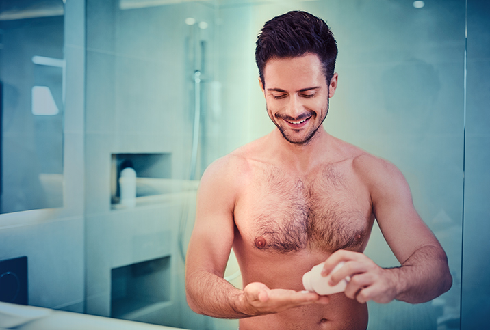 A shirtless man smiles in his bathroom while he pours a pill into his hand from a bottle.