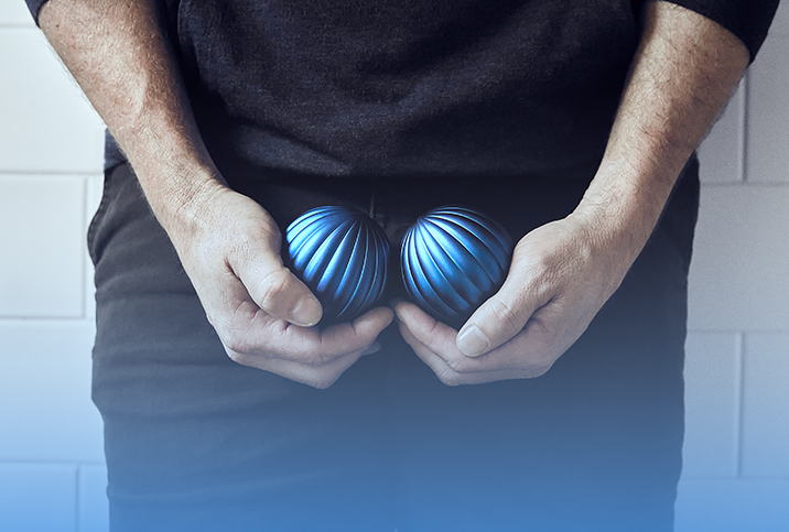 A man holds one blue ball in each hand cupped down near his groin. 