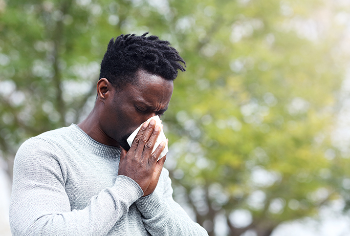 A man blows his nose into a tissue outside with a tree behind him.