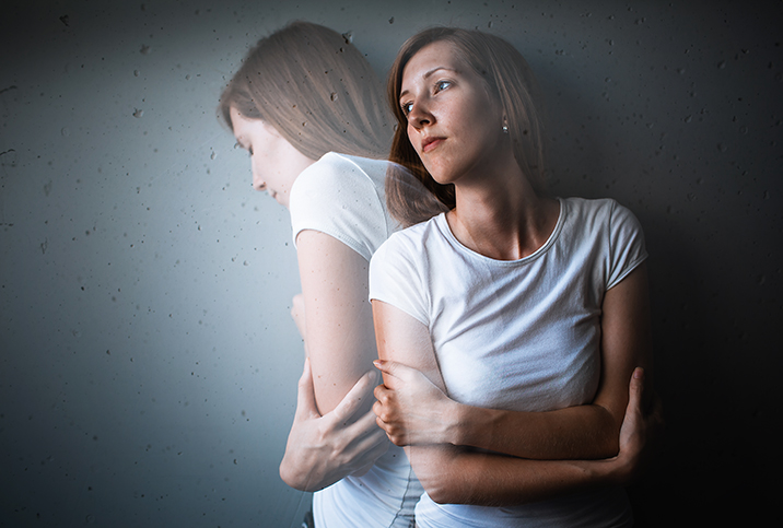 A woman leans against a wall looking to her right as a translucent image of herself turns in the same direction.