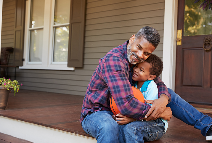 A dad sits on a front porch and hugs his child, who is holding a basketball.