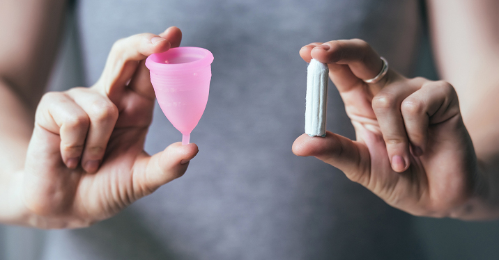 A person holds a pink menstrual cup in their right hand and an open tampon in their left.