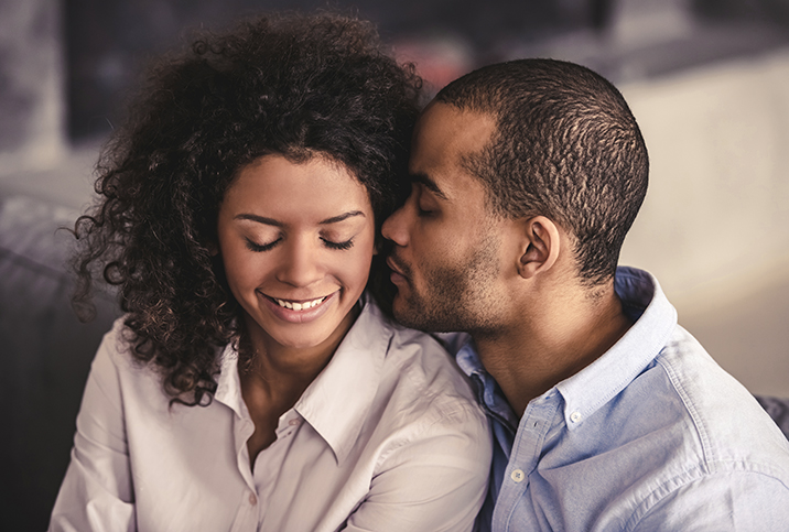 A woman closes her eyes and smiles while her partner puts his face close to her ear.