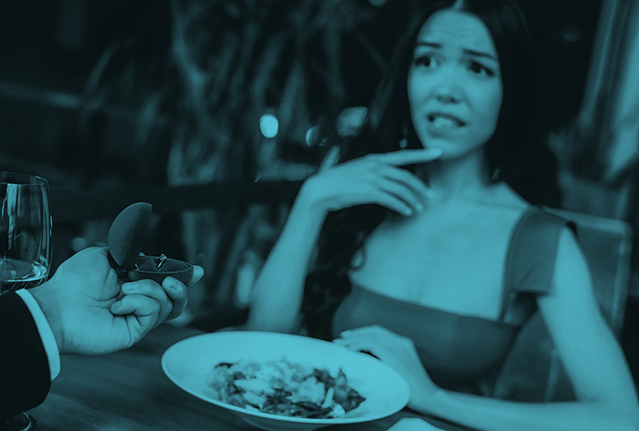 A woman looks nervously at her partner as he holds an engagement ring box in his hand over dinner.