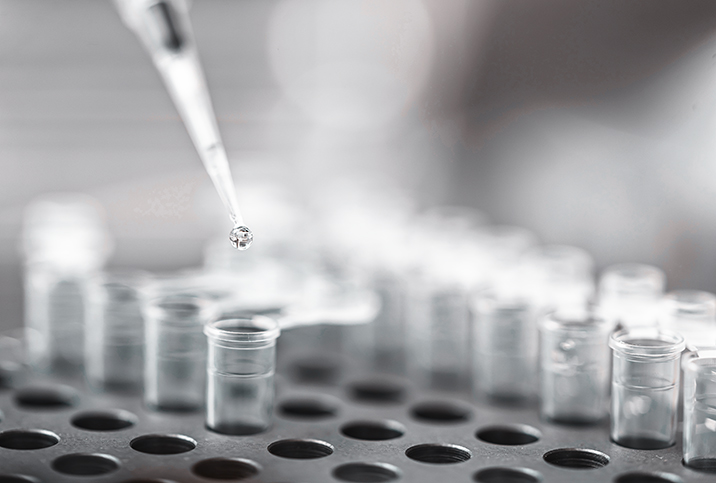 A syringe drops a liquid sample into a test tube as it sits in a rack.