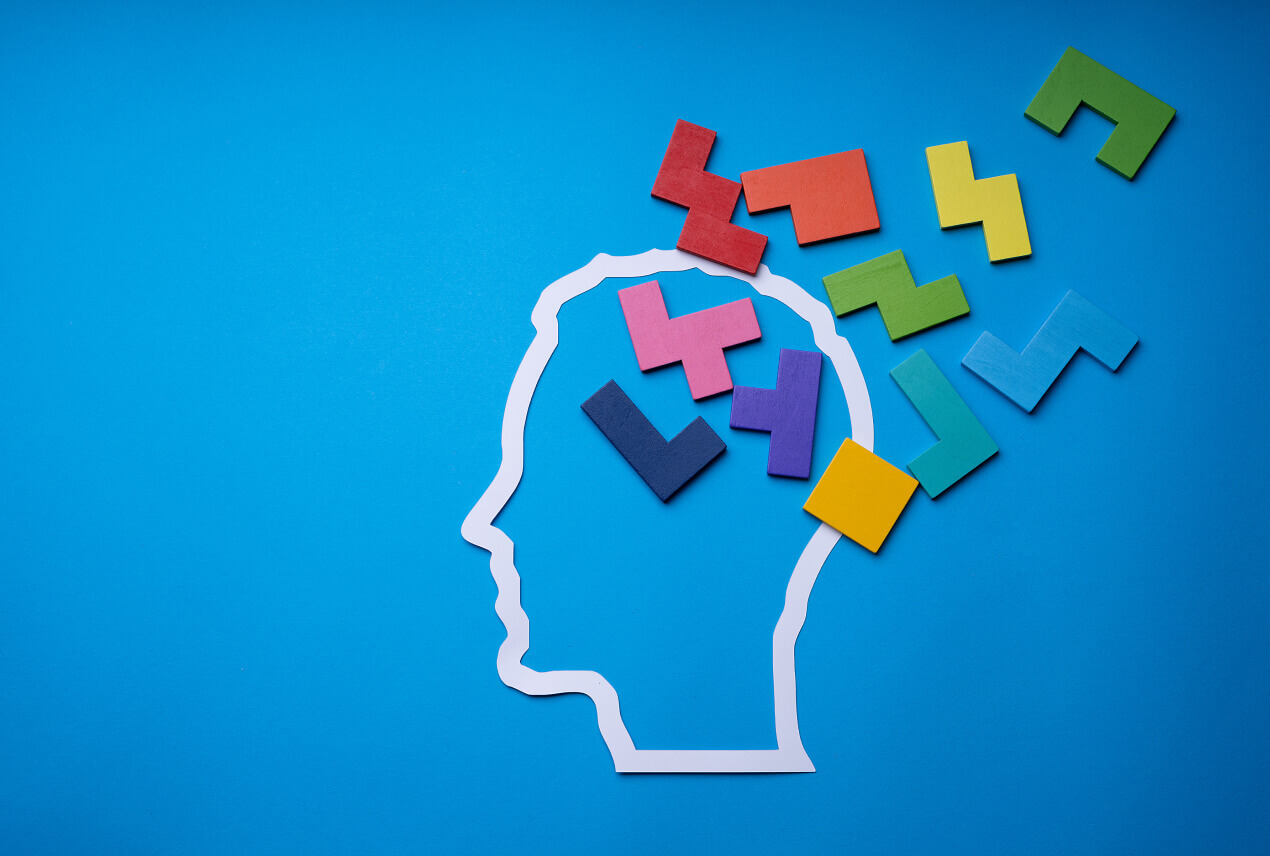 Pieces of a wooden multi-colored puzzle are coming out of the white outline of a human head against a blue background.