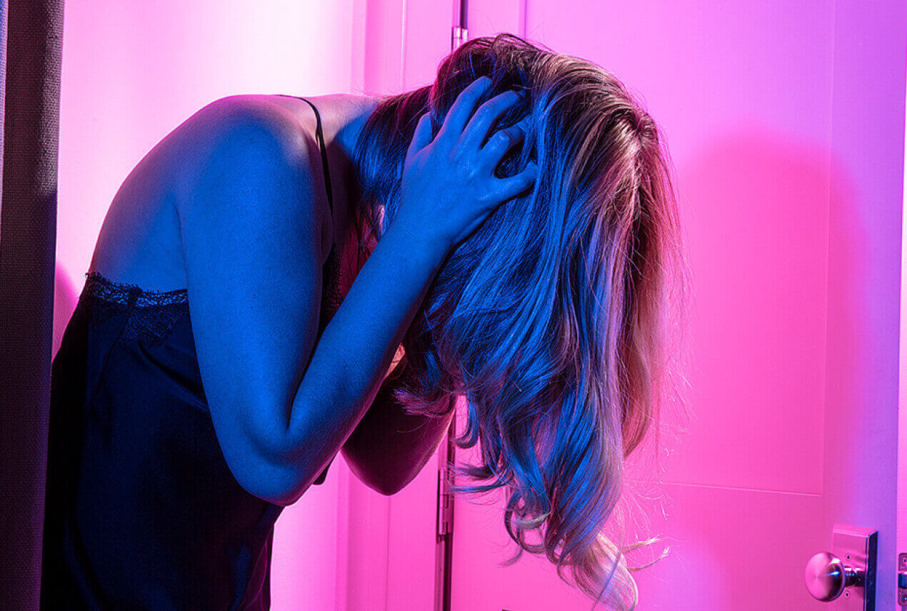 A pink light shines behind a woman who holds her head in her hands.