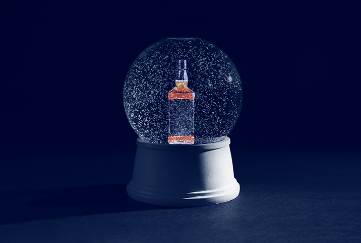 A light shines on the side of a snow globe with a bottle of whiskey inside as it sits in the dark.