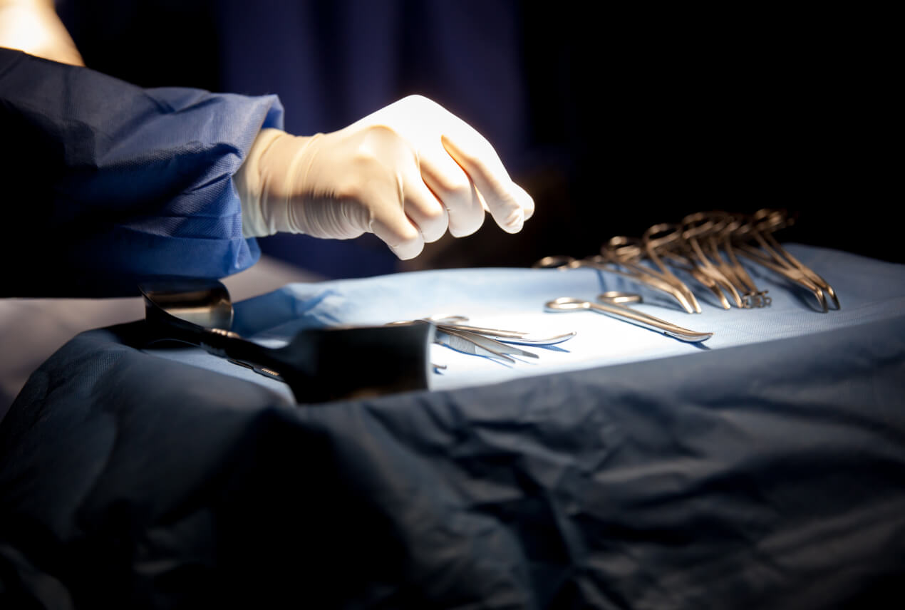 A hand covered in a surgical glove reaches for tools on a surgical tray.