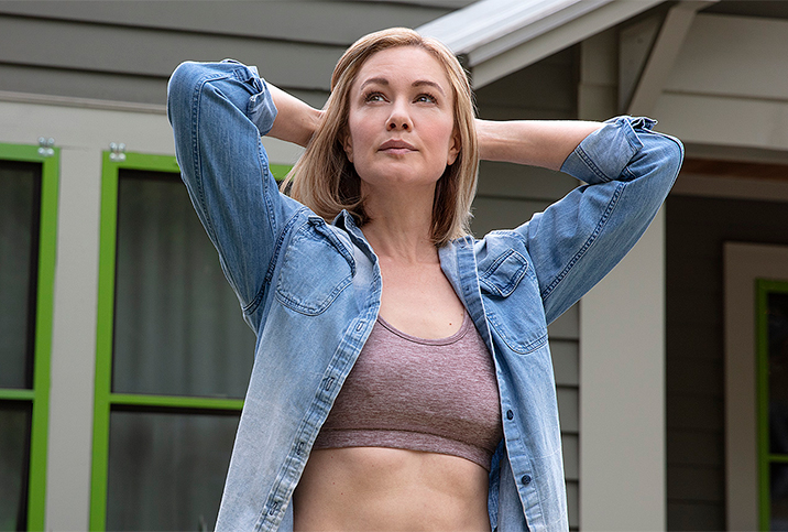 A woman in a sports bra and button down stands outside a house and holds her hands behind her head.