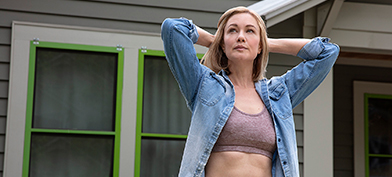 A woman in a sports bra and button down stands outside a house and holds her hands behind her head.