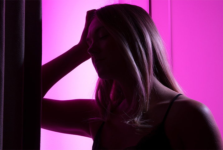 A pink light shines from behind a woman who leans her elbow against a wall and rests her head in her hands.