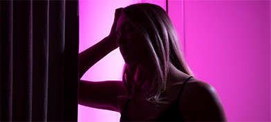 A pink light shines from behind a woman who leans her elbow against a wall and rests her head in her hands.