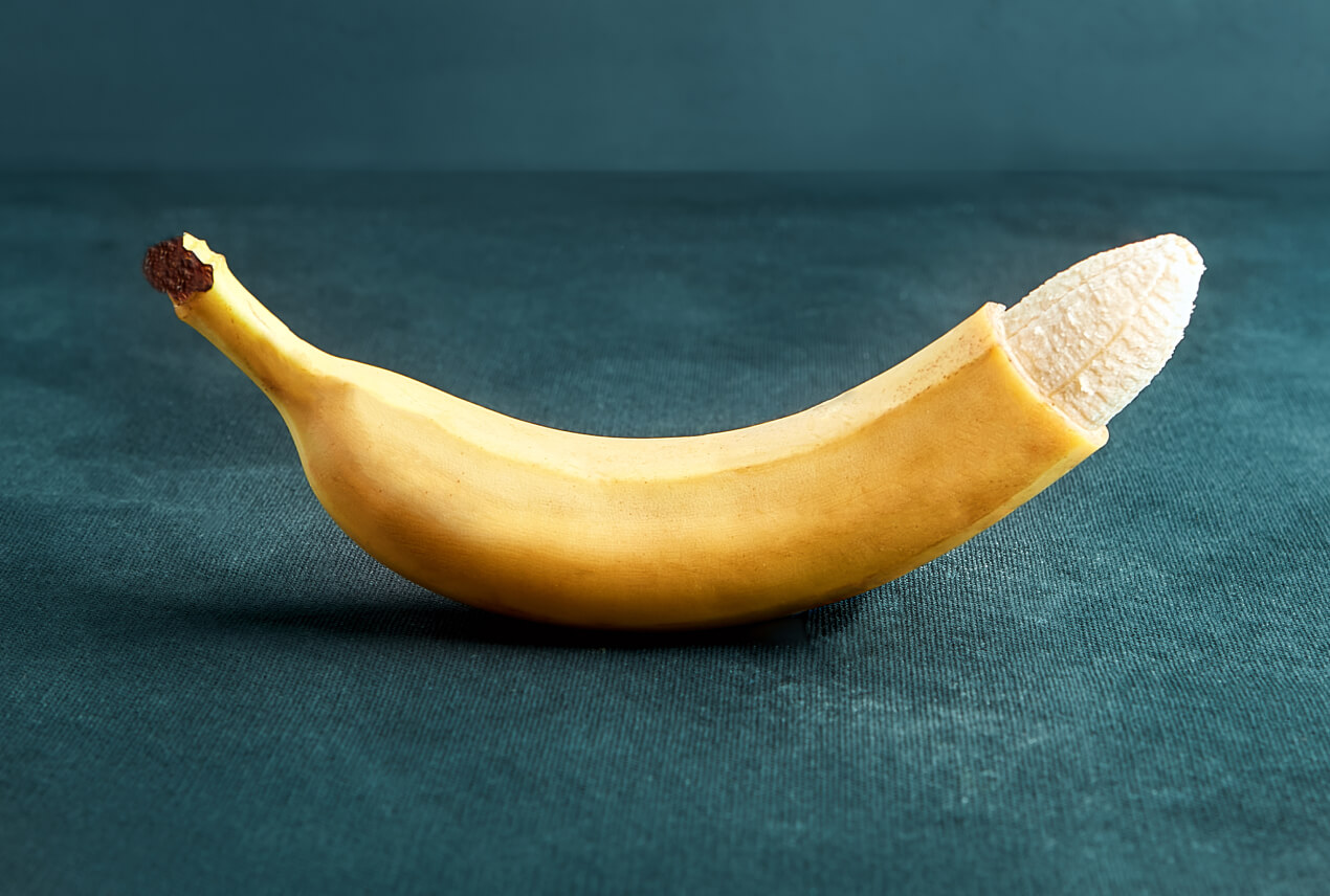 A banana sits on a dark table with the tip of the peel cut off, exposing the fruit inside.