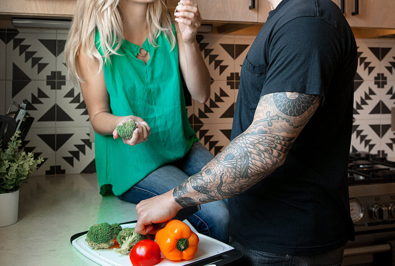A woman sits on the kitchen counter eating and a man stands by her cutting vegetables.
