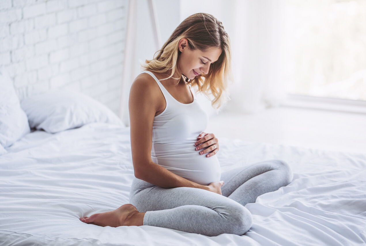 A pregnant woman holds her belly while sitting on a bed.