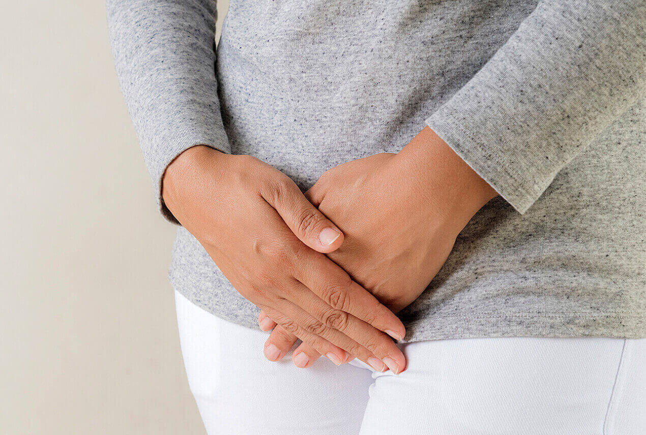 A woman in a grey sweater crosses her hands over her pelvic region