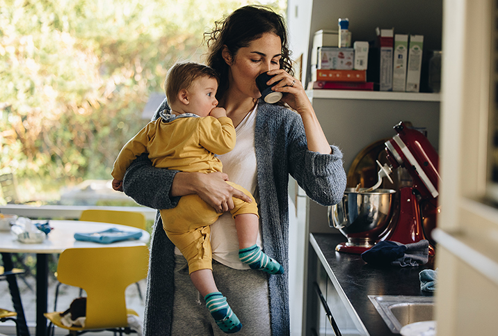 A mother holds a baby in her right arm while drinking out of a cup in her left hand.