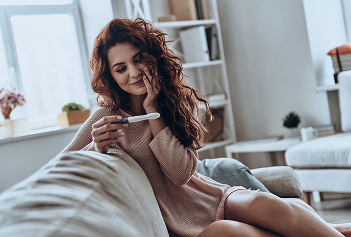 A woman sits on a sofa smiling as she holds a pregnancy test.