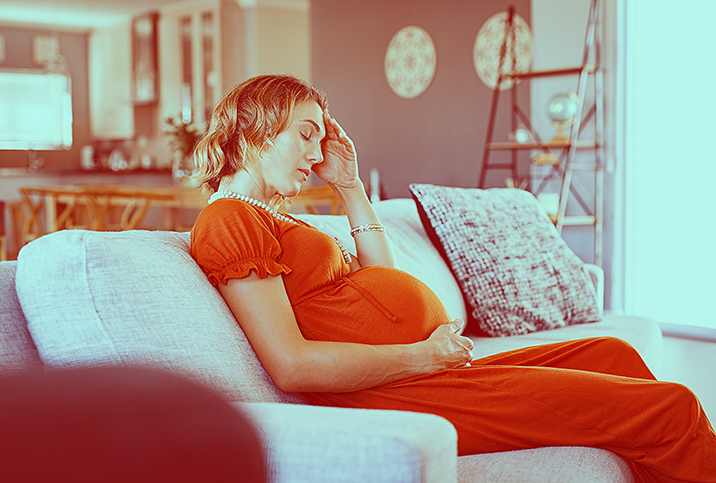 A pregnant woman sits on the sofa with her eyes closed and her hand pressed to her forehead.