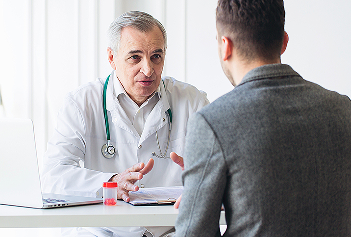 A doctor in a white coat and stethoscope gesticulates at he talks to a patient with his back to the viewer. 