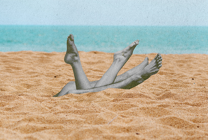 Two pairs of legs are coming out of the sand on a beach with the ocean in the background.