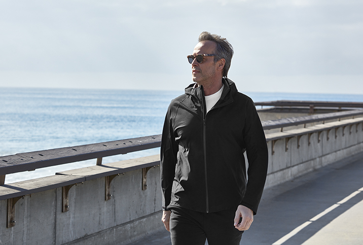 A man in activewear and sunglasses walks along a body of water in the sun.