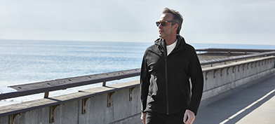 A man in activewear and sunglasses walks along a body of water in the sun.