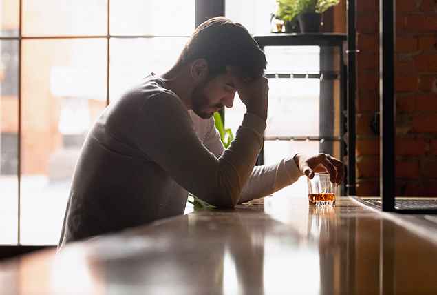 A man sits at a bar, one hand holds his head and one hand rests on a glass of alcohol.