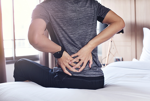 A man sits on a bed holding his lower back on the left side with both hands.