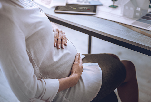A person sitting at a desk rests one hand on top and one below their pregnant belly.