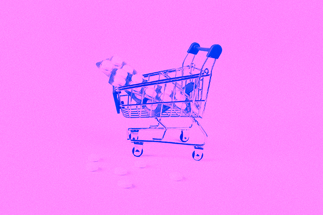 A shopping cart holds two large packets of pills against a pink background.