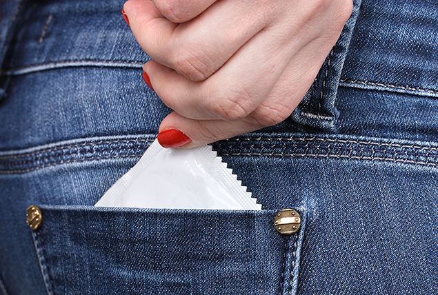 A hand with red nail polish pulls a white condom packet from a back denim pocket.