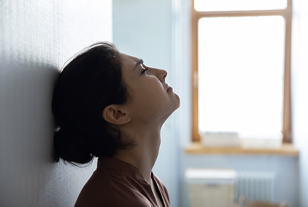A woman leans against a wall looking upwards and crying.