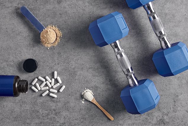 Vitamins and supplements in powder- and pill- forms lay on the ground next to two dumbbells.