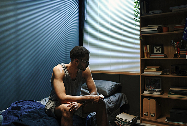 A man sits on his bed with his arms crossed along his knees.